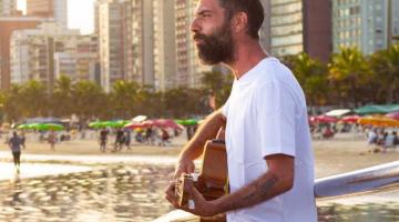 Lucas Real toca violão encostado no corrimão de ponte na praia. #Pracegover