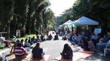 várias pessoas estão sentadas no chão, em roda, participando de uma dinâmica. Há tendas armadas no parque decoradas com balões de gás. #paratodosverem