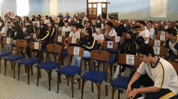 alunos sentados em auditório de escola. Há ventiladores no teto. #paratodosverem