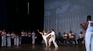 Apresentação de capoeira em palco. Jovens estão ao centro de uma roda jogando a modalidade. Ao redor, outros integrantes do grupo tocam instrumentos típicos