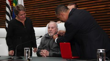 Éder Jofre está sentado na mesa da presidência. Um homem, de costas prende uma medalha na roupa do homenageado. #Pracegover