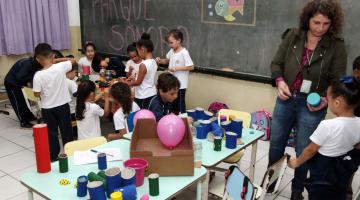 Onze crianças se reúnem em mesa com objetos reciclados. À frente, professora mostra objeto a aluna, junto a mesa com materiais coloridos