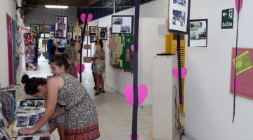 Corredor da escola está decorado com exposição de fotos e corações cor de rosa. À esquerda da imagem há uma mulher e uma criança olhando fotos que estão sobre uma mesa. #Pracegover