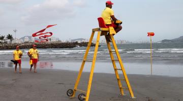 Em primeiro plano há uma escada armada na praia, com um salva vidas sentado no topo, onde há um banco. Ao fundo, outros dois guardas vêm andando. Eles estão de frente para o mar. #Pracegover