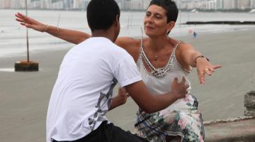 Mulher e jovem se preparam para um abraço. Eles estão sentados em uma mureta de frente para o mar. #Pracegover