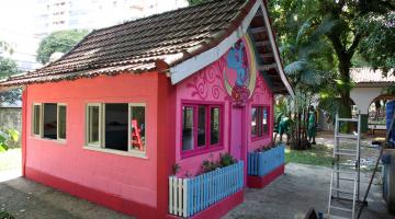 Casa de boneca com fachada frontal e lateral à vista. Lateral tem duas janelas abertas. Na frente, há duas janelas, porta e dois cercadinhos para plantas. Na frente há decoração com motivos infantis.  #Pracegover