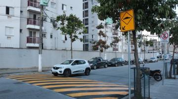 Lombada em primeiro plano com carro branco atravessando em via com prédio de fundo. #paratodosverem