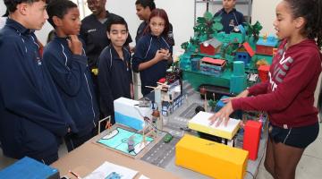 aluna está explicando maquete montada sobre mesa para outros estudantes. todos estão uniformizados. #paratodosverem 