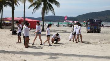 Pessoas caminham na faixa de areia carregando sacos plásticos e recolhendo lixo. #Pracegover