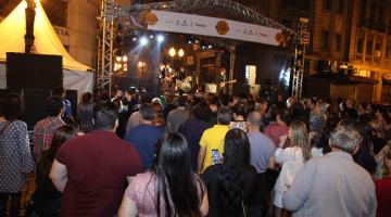 Público está na rua acompanhando show à noite. Ao fundo  há um palco com os músicos. #Pracegover