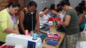 mulheres estão trabalhando em costura em torno de uma mesa. Em primeiro plano está uma máquina de costura. #paratodosverem