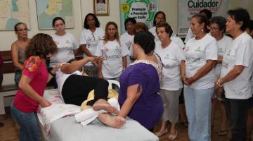 Participantes aprendem técnicas de cuidados com acamado. Uma mulher está deitada em uma cama simulando uma situação. Alunos acompanham as orientações. #Pracegover