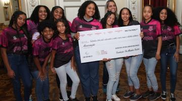 meninas posam para foto segurando cartaz que simboliza um cheque. #paratodosverem 