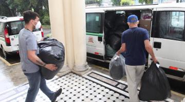 Dois homens carregam sacos com doações em direção a veículo. #paratodosverem