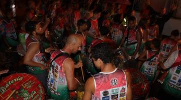 Foliões com camisetas iguais estão em rua com bateria. #Pracegover 
