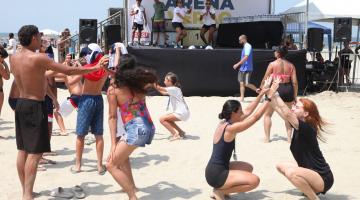 pessoas dançam e se movimentam na faixa de areia diante de um palco em cujo fundo se lê Arena. #paratodosverem 