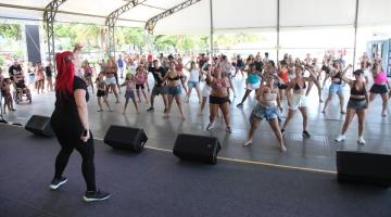 professora dá aula para grande grupo de pessoas, predominantemente mulheres, sob tenda. Ela está no palco, em primeiro plano. O publico se movimente, de frente para a foto, ao fundo. #paratodosverem 