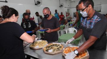 Uma pessoa atrás de uma mesa , onde estão três pizzas , algumas pessoas na fila para comer #paratodosverem