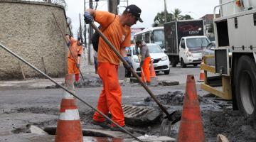 Homem remove lama de bueiro em rua. Há cones delimitando a área de liimpeza