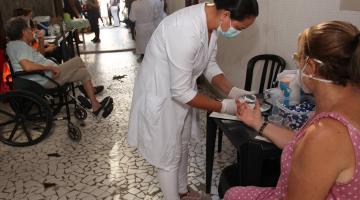 mulher usando avental branco tira gota de sangue da ponta do dedo de outra mulher para teste de glicemia. #paratodosverem