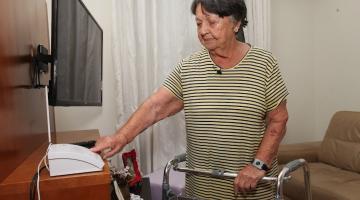 idosa com andador aponta para telefone na sala de seu apartamento #paratodosverem