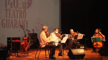 quatro músicos de cordas estão sentados se apresentado num palco. Ao fundo se lê 140 anos Teatro Guarany. #paratodosverem 