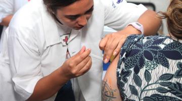 mulher usando avental branco vacina braço esquerdo de mulher que está sentada e de costas para a foto. #paratodosverem 
