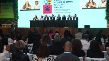 palco com mesa e pessoas sentadas. Ao fundo um telão com imagens. Público está sentado, em primeiro plano, assistindo às apresentações. #paratodosverem 