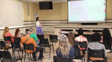 salão com pessoas sentadas assistindo uma palestra. Ao fundo uma mulher em pé fala ao grupo. #paratodosverem