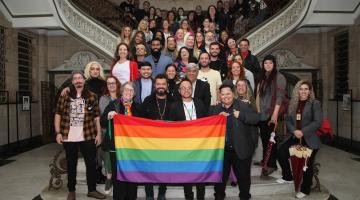 integrantes de conselho posam para foto na escadaria do saguão da prefeitura. à frente, a bandeira lgbt.#paratodosverem