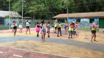 mulheres estão dançando em quadra com grande vegetação ao fundo. #paratodosverem