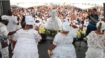 mulheres vestidas de branco estão em palco de costas para a foto. Imagem de iemanjá está à frente com o público ao fundo. #paratodosverem