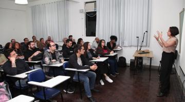 mulher está em pé falando a um grupo de pessoas sentadas. #paratodosverem 