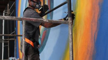homem faz grafite em muro #pracegover 