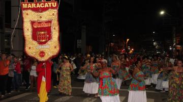 cortejo do grupo maracatu quiloa por rua. Homem à frente segura estandarte e mulheres vem dançado com vestidos coloridos. #paratodosverem