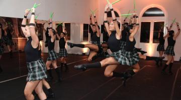 Bailarinas dão salatos no ar. Todas estão seguram um tipo de baqueta em cada mão, ambas levantadas para o alto. #paratodosverem 