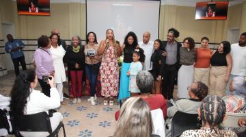 mulheres e homens reunidos em salão. Uma delas posa com troféu. #paratodosverem