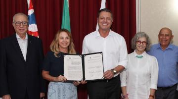 cinco pessoas posam para a foto. Ao centro, o prefeito e a secretária de educação seguram pasta com documento assinado. #paratodosverem 