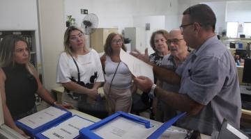 pessoas estão reunidas em um escritório. Um deles mexe em um papel e mostra para os outros. #paratodosverem 