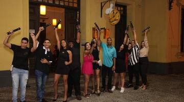 jovens estão na porta do restaurantes segurando seus canudos de formatura com as mãos para o alto, em tom de comemoração. #paratodosverem