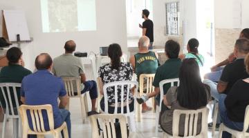 pessoas estão sentadas em sala assistindo a uma apresentação em um telão. #paratodosverem