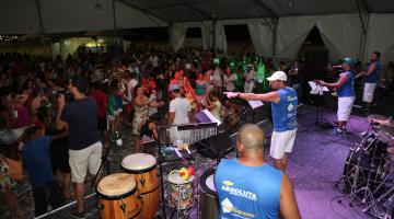 Baile de carnaval em tenda. No palco, os músicos se movimentam. Há instrumentos de percussão. #Paratodosverem