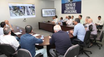 pessoas sentadas em torno de uma mesa assistindo a uma apresentação em um telão ao fundo. #paratodosverem 