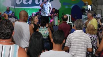 Happy Hour em tenda com cantor ao fundo. Atrás dele há um painel onde se lê Santos Verão. À frente do cantor e de costas para a foto está o  público. #Pracegover