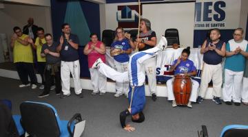 capoeirista estão de cabeça baixo, com as mãos no chão e de pernas para o ar. Atrás, o grupo acompanha com palmas, berimbau e atabaque. #paratodosverem