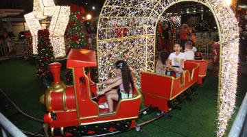 trenzinho circula por trilhos e passa sob túnel de luzes. Crianças dentro do brinquedo sorriem. #paratodosverem 