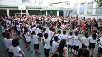 Crianças estão alinhadas em pátio de frente para as mães #pracegover 