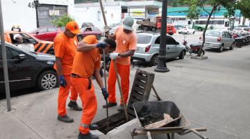 Funcionários uniformizados, com luvas e instrumentos de cabos longos para inserir na rede de drenagem fazem a limpeza em bueiro de calçada. #Pracegover