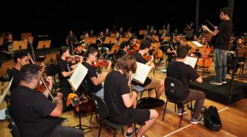 Jovens músicos estão sentados e tocando seus instrumentos. O maestro está à frente do grupo, sobre um degrau. #Pracegover