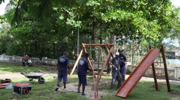 Homens estão sobre gramado de praça mexendo em peças de madeira para formar um balancê. #Pracegover